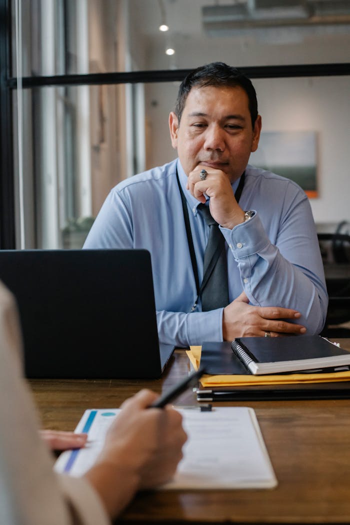 about-us-02 Serious mature ethnic entrepreneur sitting at wooden table with documents and laptop and thinking while crop anonymous colleague taking notes on clipboard