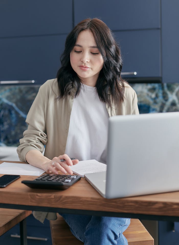 heros-img A young woman calculating finances at home using a laptop and calculator, reflecting a focused budgeting process.
